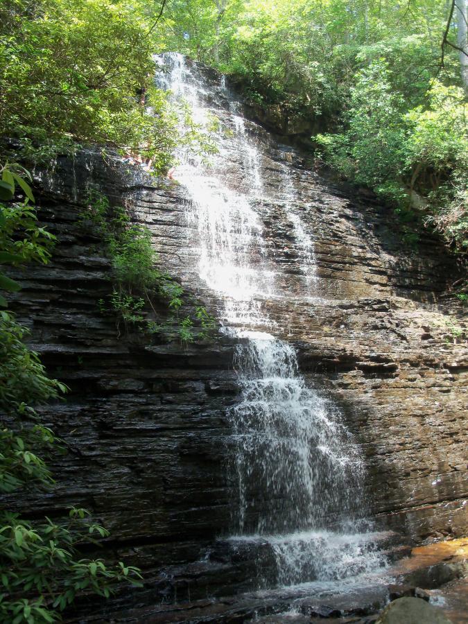 A cascading waterfall flowing down layered rock formations, surrounded by lush green foliage. Sunlight filters through the trees, creating a serene and tranquil natural setting. Chilhowee trail system mountain bike trail.