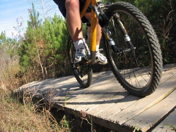 A close-up view of a mountain bike riding on a wooden plank path surrounded by greenery, with a focus on the bike's front wheel and the rider's leg. Blankets Creek mountain bike trail.