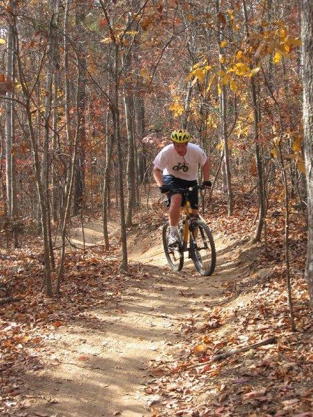 A person riding a mountain bike on a narrow dirt trail surrounded by tall trees with autumn leaves. The scene captures the essence of outdoor biking in nature, showcasing the vibrant fall foliage in the background. The cyclist is wearing a helmet and riding with focus along the winding path. Blankets Creek mountain bike trail.