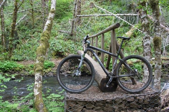 A mountain bike is leaning against a wooden bridge in a lush forest setting, with a small stream visible in the background. The bike is black and has visible dirt, suggesting recent use. Surrounding the scene are vibrant green foliage and trees. Tillamook State Forest mountain bike trail.
