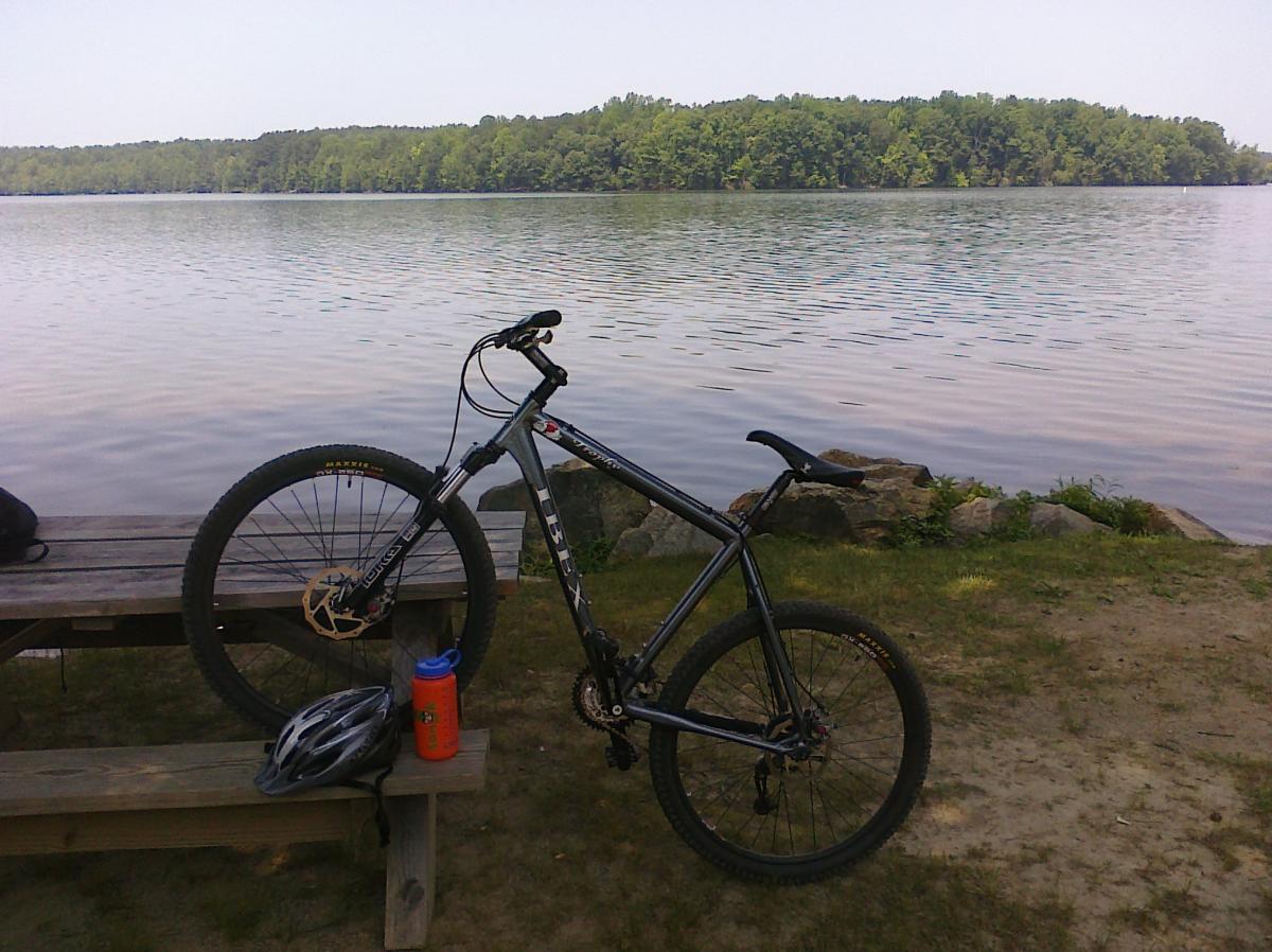 A mountain bike rests on a wooden picnic table near a calm lake. The scene includes a water bottle and a helmet placed on the table, with trees lining the opposite shore of the water. The sky is clear, suggesting a sunny day. Wild Turkey mountain bike trail.