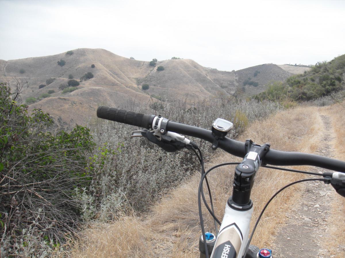 A mountain bike handlebar is shown in the foreground, with a view of rolling, dry hills and sparse vegetation in the background. The scene is set on a dirt trail surrounded by brush and grass, under a cloudy sky. New Millennium Loop mountain bike trail.