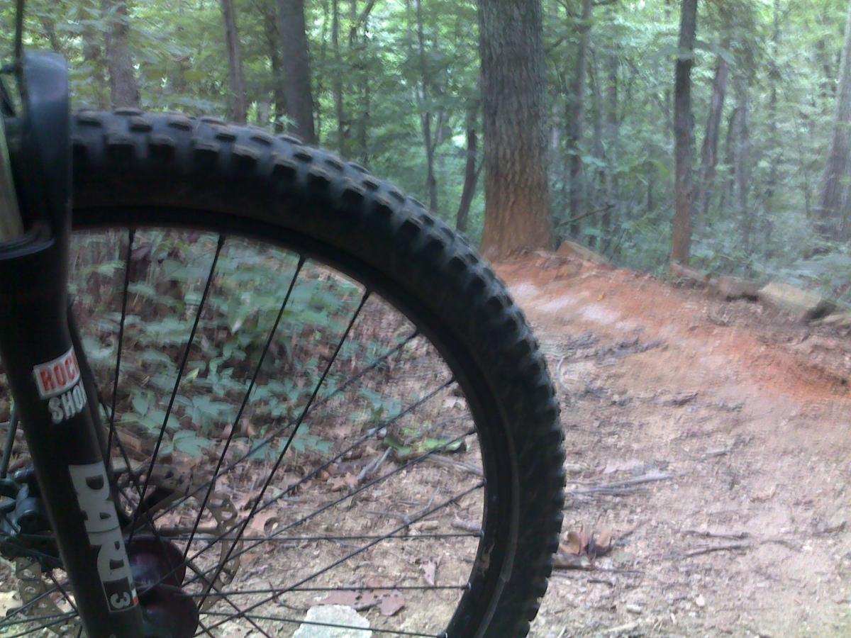 Close-up of a mountain bike wheel on a dirt trail in a forested area, with trees and greenery in the background. The trail appears rugged, indicating potential for off-road cycling. Hobby Park mountain bike trail.