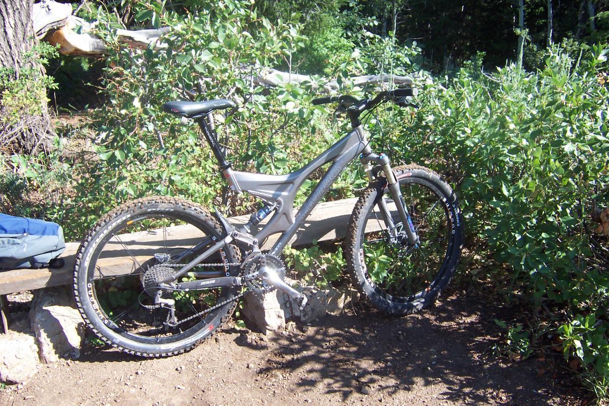 A silver mountain bike is parked beside a wooden bench in a natural outdoor setting, surrounded by lush green foliage and trees. The bike features thick, knobby tires suitable for rugged terrain and has visible gears and a suspension fork, suggesting it is designed for off-road riding. Jardine Juniper mountain bike trail.