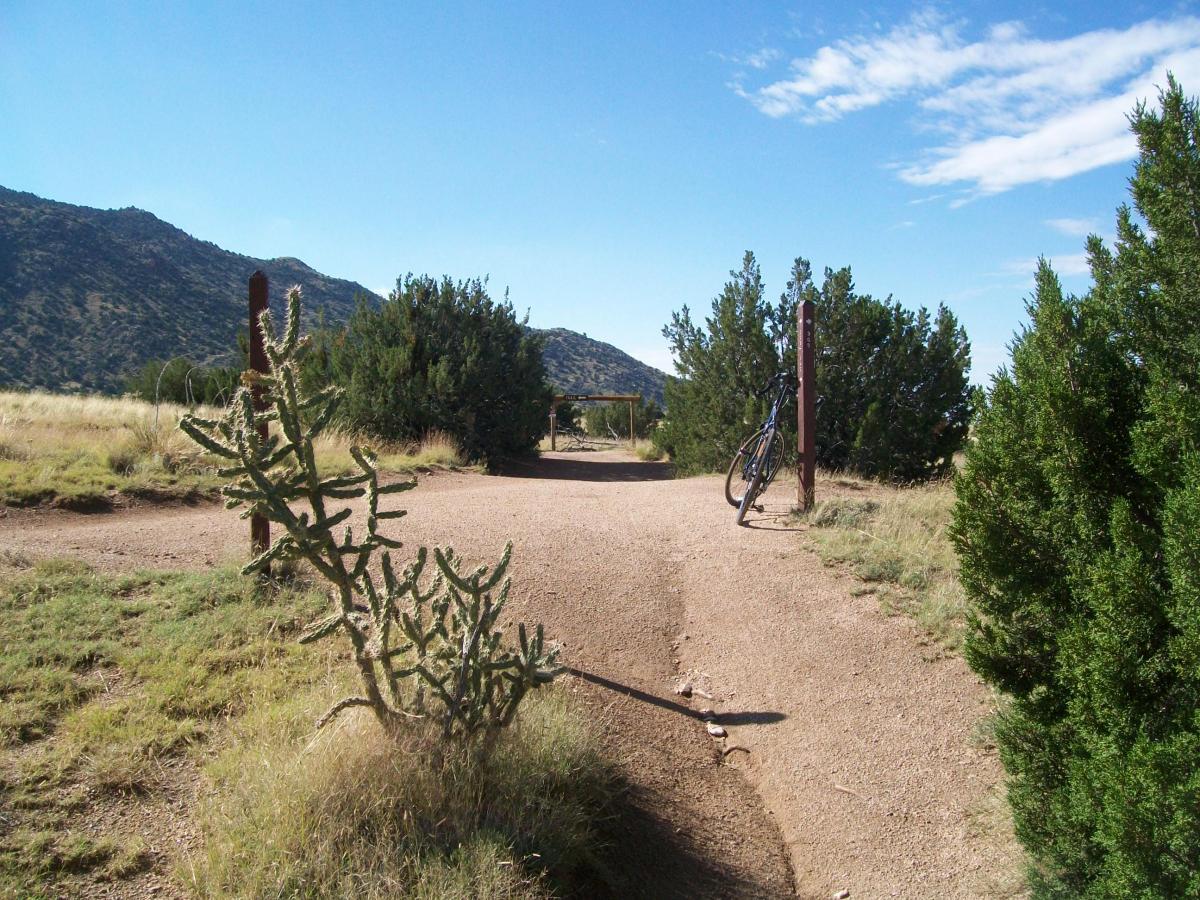 A dirt path winding through a semi-arid landscape, flanked by shrubs and cacti, with a mountain range in the background under a clear blue sky. A bicycle is leaning against a post near the path's entrance. South Foothills mountain bike trail.