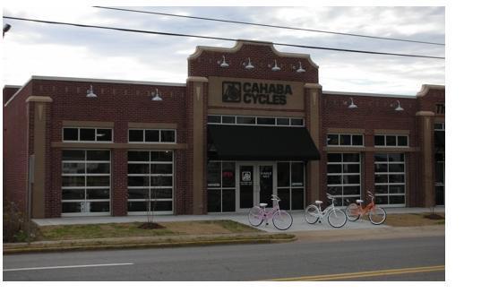 Front view of a brick building featuring the sign "Cahaba Cycles," with large glass windows and a black awning. Several bikes in pastel colors are parked in front, and the scene is set on a street with cloudy skies above.