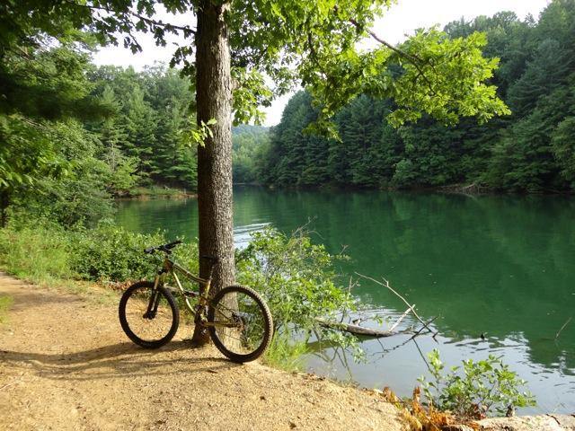 A mountain bike leaning against a tree beside a calm, green lake surrounded by lush forest. The scene captures a tranquil outdoor setting with a dirt path leading along the water's edge. Dark Mountain Trail mountain bike trail.