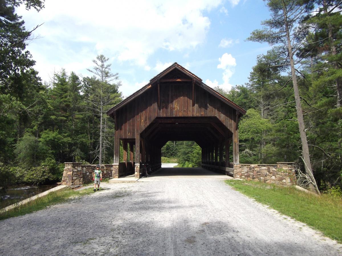 A wooden covered bridge stands in the center of the image, framed by tall trees under a clear blue sky. A gravel path leads under the bridge, while a stone wall is visible on either side. A person, dressed in colorful clothing, walks along the path near the entrance of the bridge. DuPont State Forest mountain bike trail.