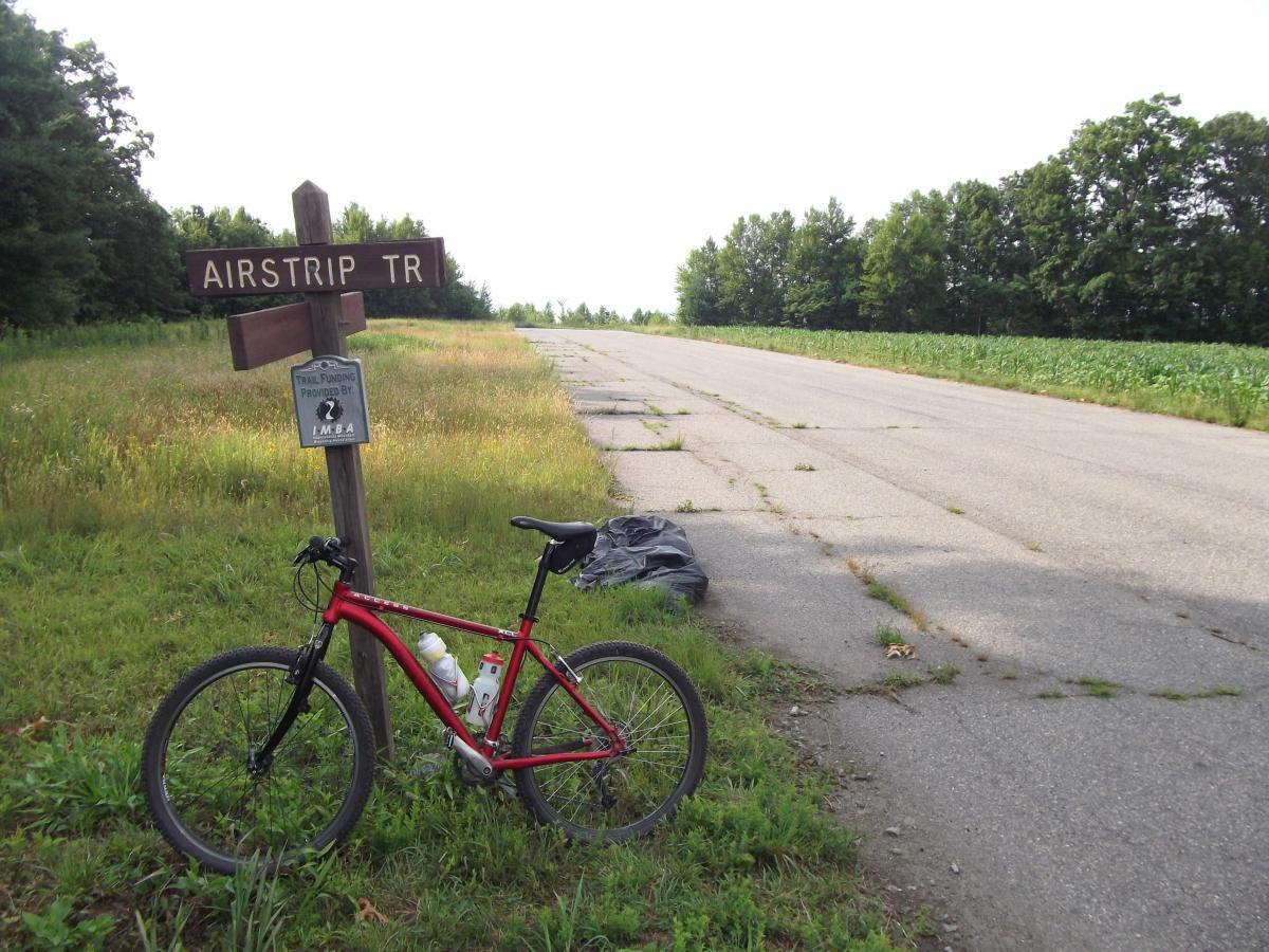 A red bicycle is parked next to a wooden signpost indicating "AIRSTRIP TR" along a gravel road. In the background, lush greenery and a field can be seen, with a few scattered clouds in the sky. A trail maintenance sign is attached below the main signpost, and a black bag is positioned on the grass beside the bike. DuPont State Forest mountain bike trail.