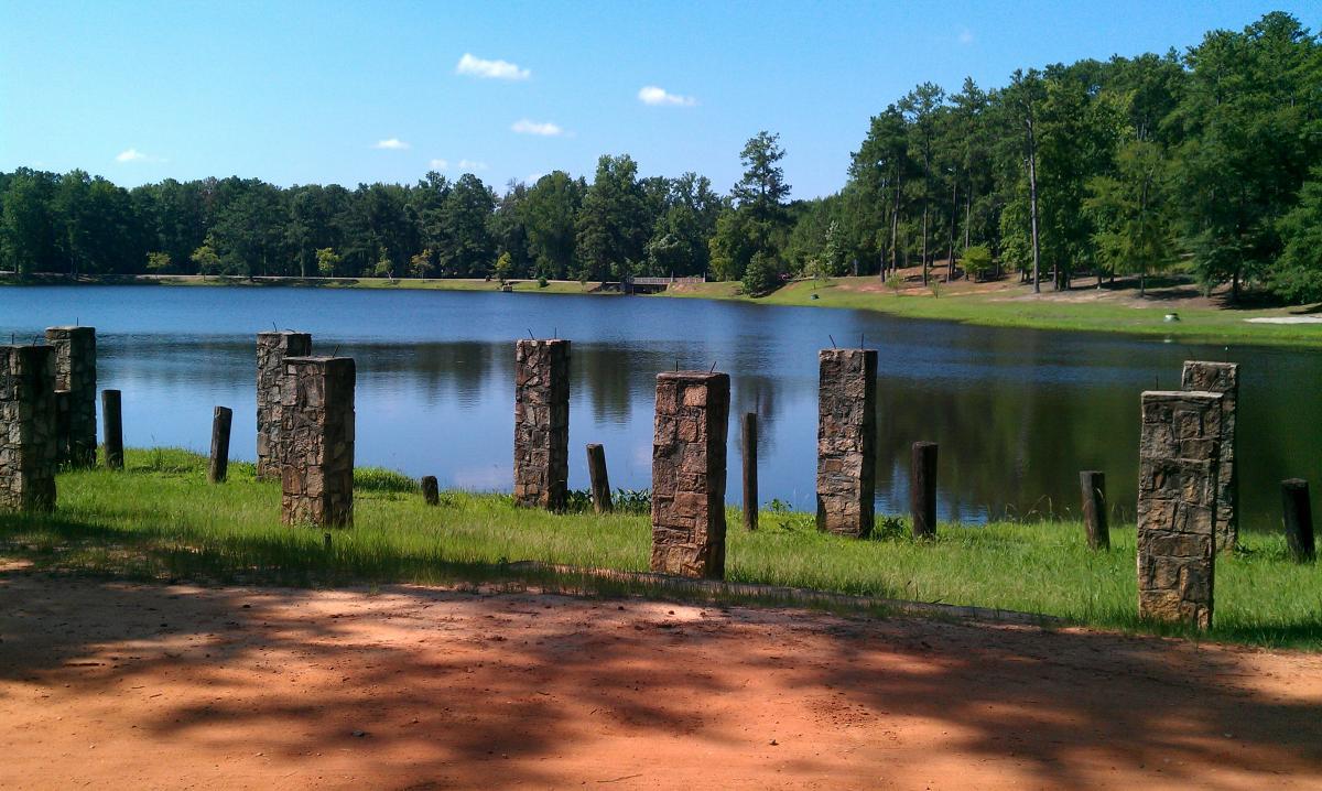 A serene lake surrounded by greenery, featuring stone pillars and wooden posts in the foreground. The calm water reflects the blue sky and scattered clouds, with trees lining the opposite shore. The scene conveys a tranquil outdoor setting. Flat Rock Park mountain bike trail.