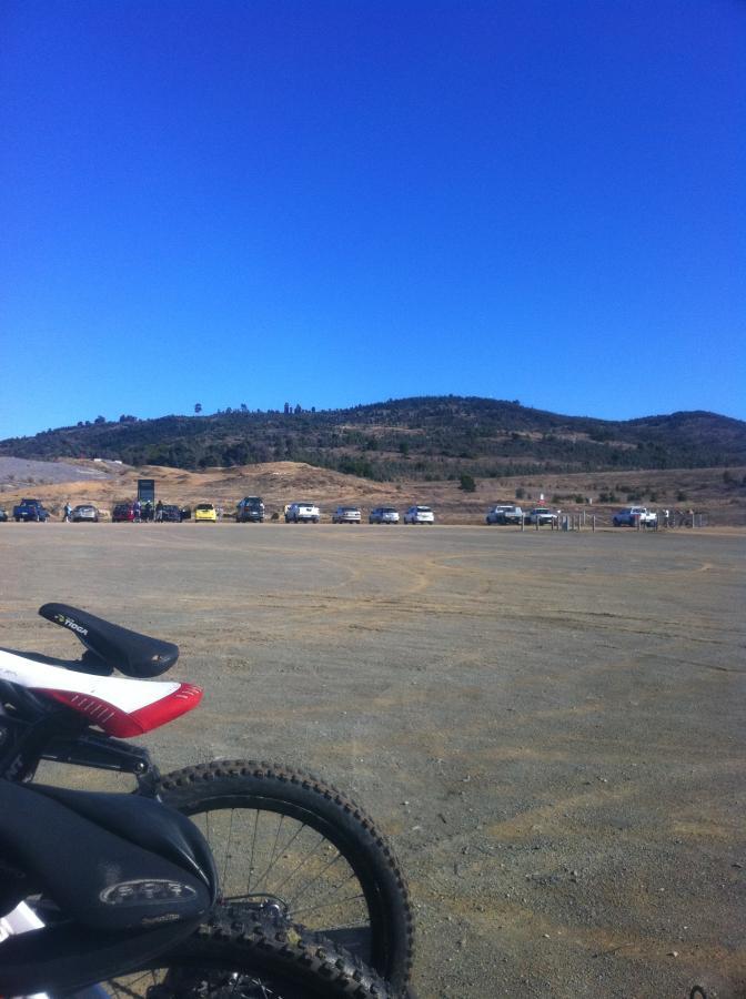 A dirt parking area with several cars lined up in the background, a mountainous landscape under a clear blue sky, and the front wheel of a mountain bike in the foreground. Stromlo Forest Park mountain bike trail.