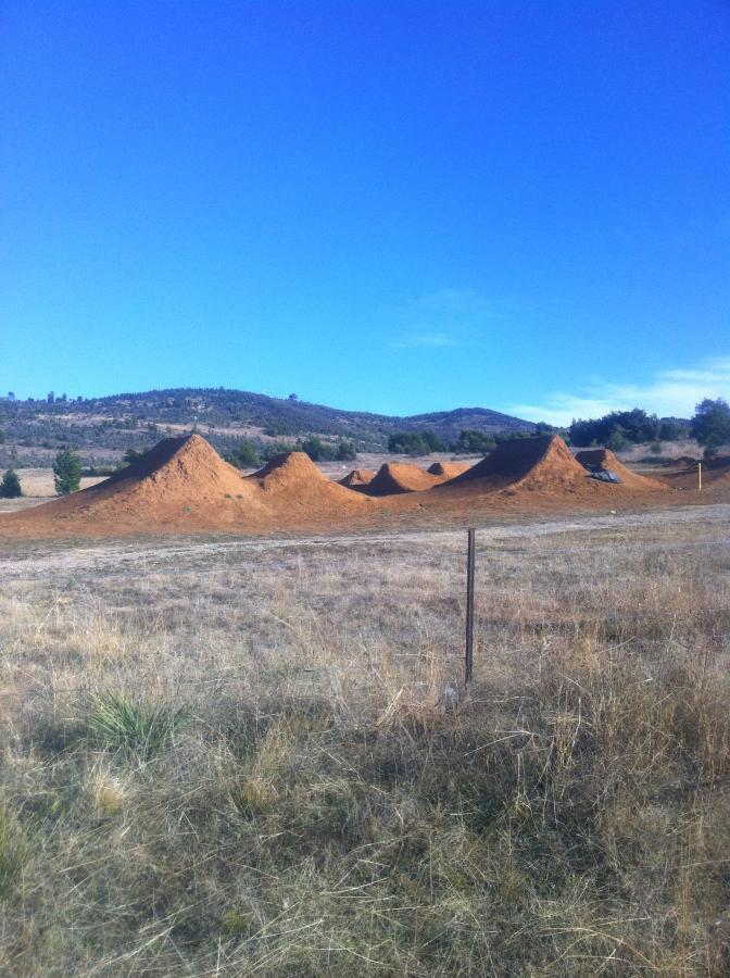 A landscape featuring several dirt jumps or mounds in a grassy area, with rolling hills in the background under a clear blue sky. A barbed wire fence lines the foreground, and sparse trees can be seen beyond the jumps. Stromlo Forest Park mountain bike trail.
