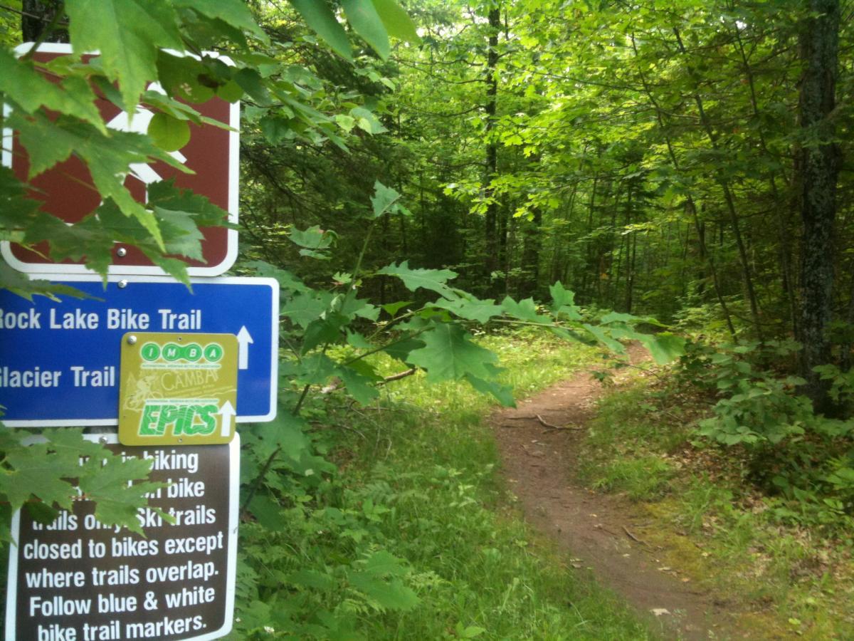 Trail signs indicating the Rock Lake Bike Trail and Glacier Trail, surrounded by dense green foliage and trees. The path ahead winds through the forest, suggesting an inviting biking route. Rock Lake mountain bike trail.