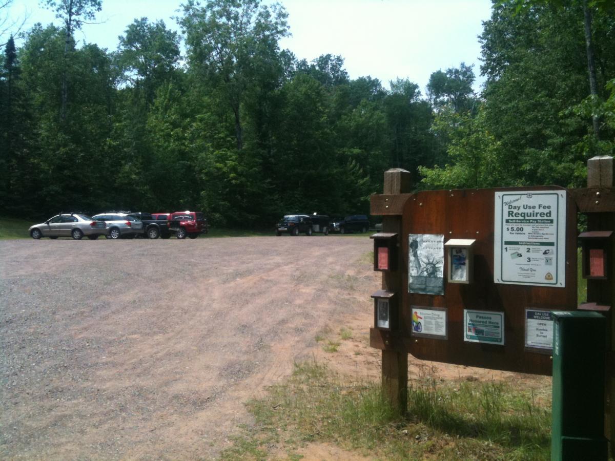 Image of a gravel parking lot surrounded by trees, with several parked cars. A wooden signpost in the foreground displays information about day use fees, including pricing and other guidelines for visitors. The scene is set on a clear, sunny day. Rock Lake mountain bike trail.
