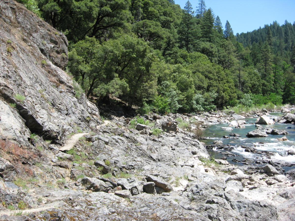 A rocky riverbank surrounded by lush greenery, with a small path winding alongside the water. The scene features a mountainous backdrop, clear blue skies, and a flowing river with scattered boulders. Old Flume Trail / South Yuba Trail mountain bike trail.