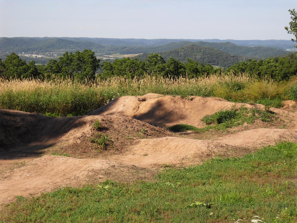 A scenic landscape view featuring rolling hills and valleys under a clear blue sky. In the foreground, there is a dirt terrain with mounds and gentle slopes, surrounded by a mix of grass and wild plants. The background showcases distant hills and a patch of woodlands, highlighting the natural beauty of the outdoors. Hixon Forest mountain bike trail.