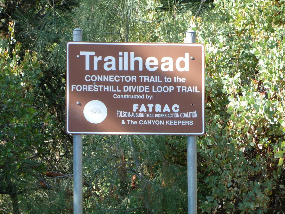 Alt text: A brown trailhead sign indicating the connector trail to the Foresthill Divide Loop Trail, constructed by FATRAC (Folsom-Auburn Trail Riders Action Coalition) and The Canyon Keepers, with greenery in the background. Clementine / Forresthill Connector Trail mountain bike trail.