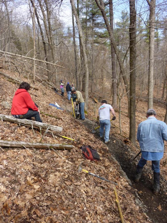 A group of people working on a hillside in a wooded area, using tools like shovels and picks. The ground is covered with dry leaves, and some individuals are engaged in digging or clearing while others are resting or observing. The scene shows a mix of trees, some still bare of leaves, indicating early spring or late winter. Lippman Park mountain bike trail.