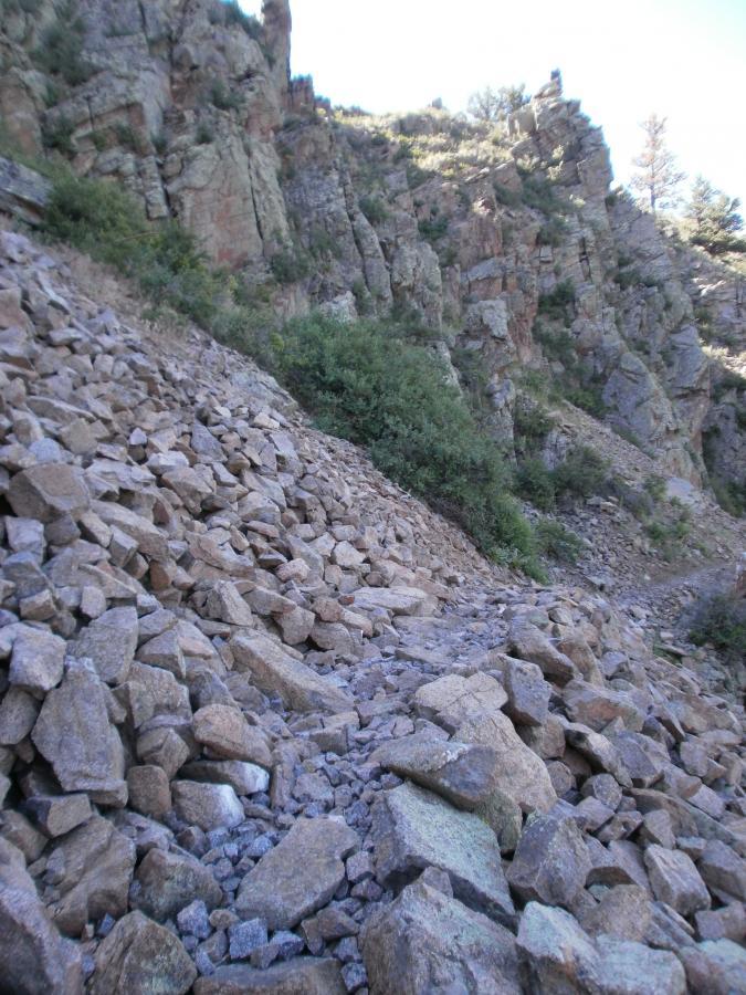 Rocky hillside with loose stones and boulders scattered on a sloped surface, surrounded by sparse vegetation and steep rock formations in the background. Curt Gowdy State Park mountain bike trail.
