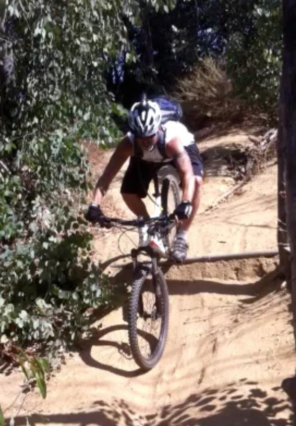 A mountain biker navigating a sandy trail, leaning forward as they skillfully maneuver their bike over a small incline surrounded by greenery. Fullerton Loop mountain bike trail.
