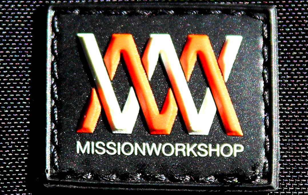 Logo patch for Mission Workshop featuring a stylized letter design in orange and white on a black background, with the text "MISSIONWORKSHOP" displayed beneath the graphic.
