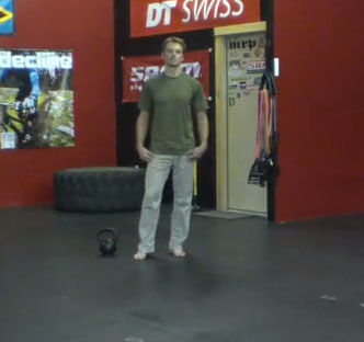 A person standing in a gym or fitness studio, wearing a green t-shirt and light-colored pants, positioned barefoot on a black rubber floor. A kettlebell is placed on the floor nearby, and the background features various fitness-related posters and equipment.