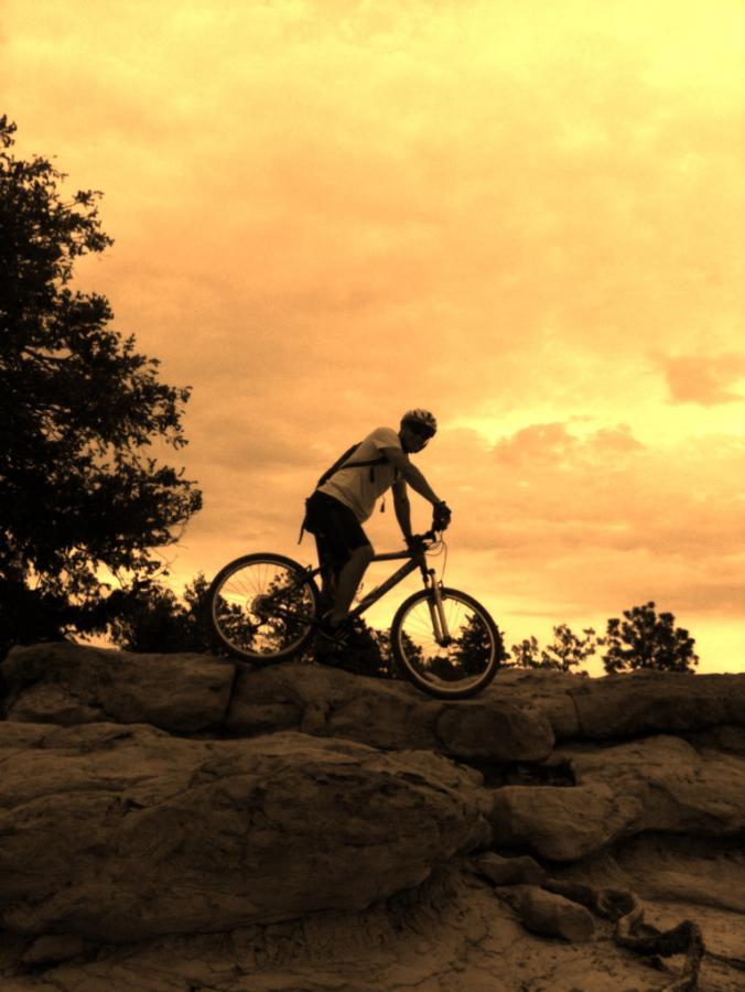 A silhouette of a person on a mountain bike, poised on a rocky surface with trees in the background, against a dramatic orange and yellow sky. Ute Valley Park mountain bike trail.