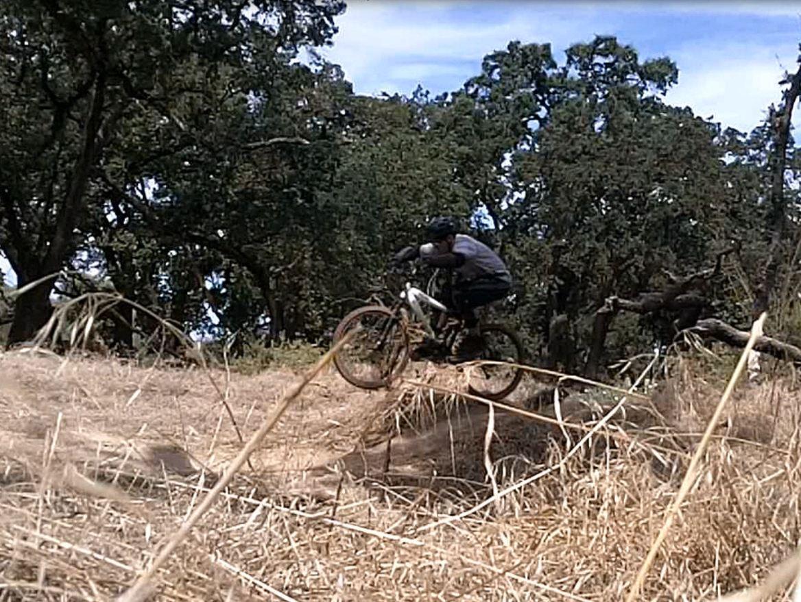 A mountain biker jumps over a small mound of dirt surrounded by dry grass and trees in a wooded area on a sunny day. Ripon Riding Spot mountain bike trail.