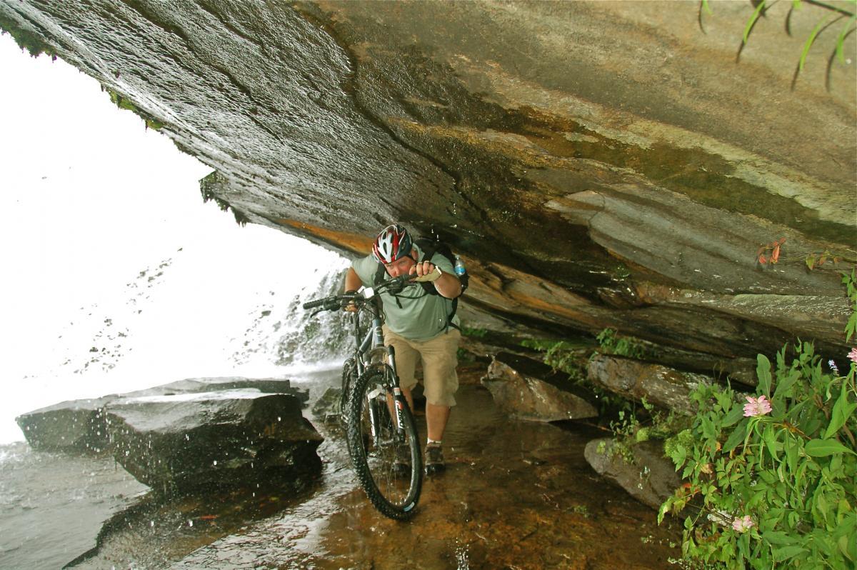 A mountain biker navigating through a rocky, wet area beneath a large overhanging rock, with water cascading in the background. The rider, wearing a helmet and casual outdoor clothing, is leaning forward, trying to maintain balance while pushing the bike. Surrounding vegetation includes ferns and small flowers, adding a touch of nature to the scene. DuPont State Forest mountain bike trail.