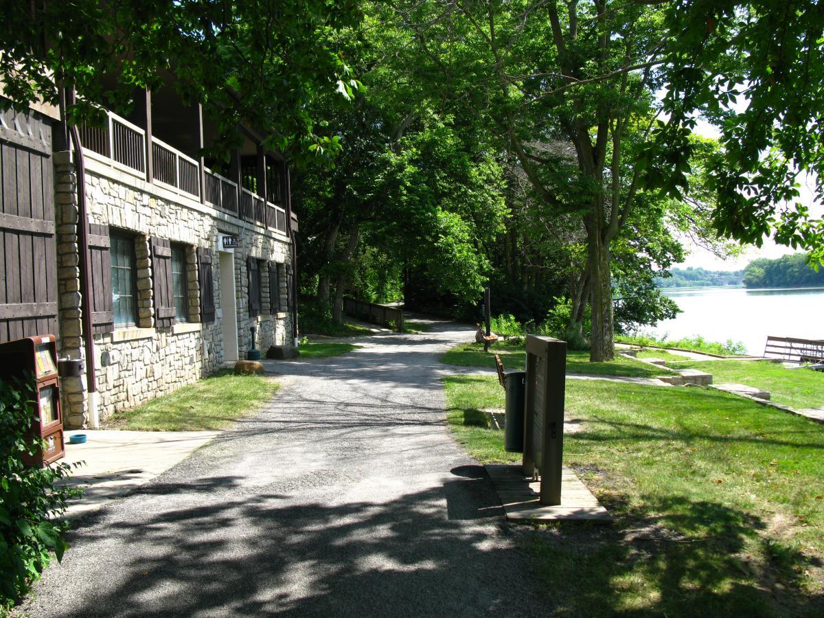 A scenic pathway next to a stone building, surrounded by lush green trees, leading towards a river. The path features grassy areas, a garbage can, and a distant view of the water, creating a serene outdoor atmosphere. Farnsworth mountain bike trail.