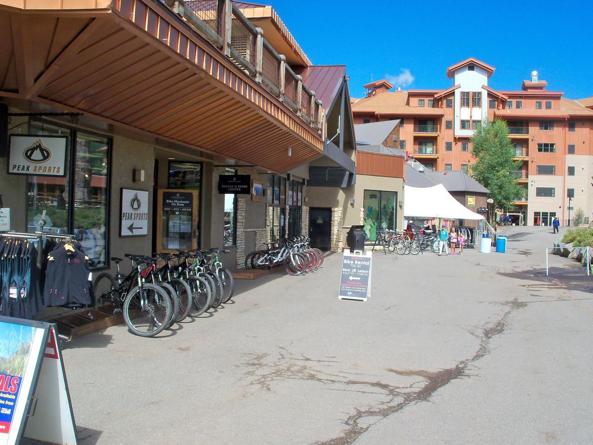 A storefront of a sports rental shop called "Peak Sports," featuring a row of bicycles outside. The image shows a paved walkway with a few people walking nearby and a large building in the background. There are signs promoting bike rentals and nearby shops. The scene is set under a clear blue sky. Evolution Bike Park at Crested Butte Mountain Resort mountain bike trail.