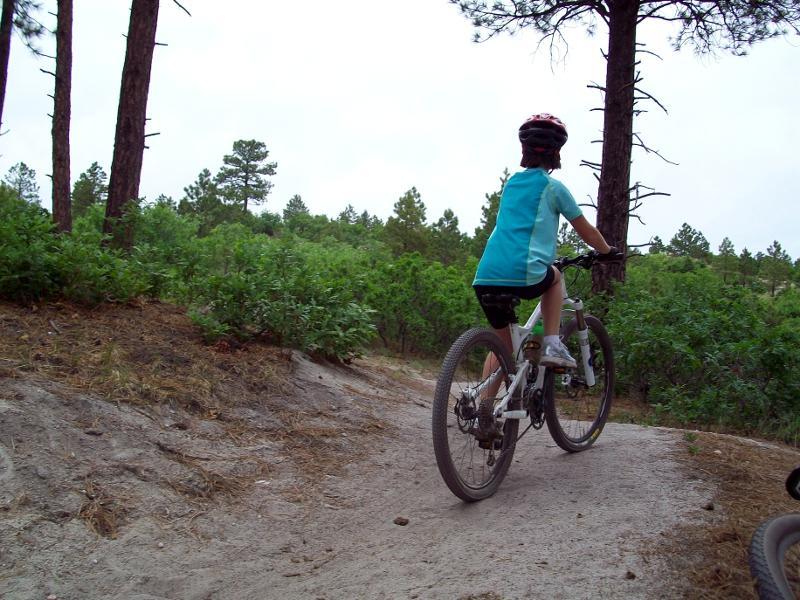 A child riding a mountain bike on a dirt trail surrounded by trees and greenery, viewed from behind. The child is wearing a blue shirt and a helmet, navigating a winding path through a natural environment. Dawson's Butte mountain bike trail.
