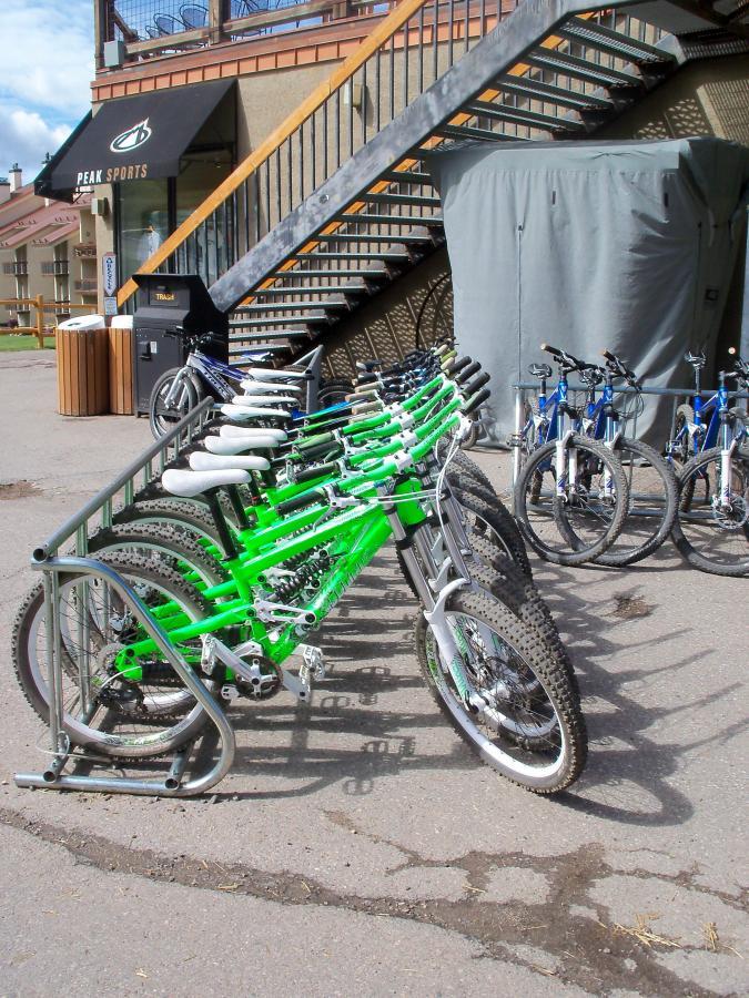 A row of bicycles, primarily in bright green, is lined up in a bike rack outside a building. In the background, there are additional bikes in blue. A set of stairs leads up to the entrance of a business labeled “Peak Sports,” with a covered area visible. The scene is set in a paved outdoor space under a clear sky. Evolution Bike Park at Crested Butte Mountain Resort mountain bike trail.