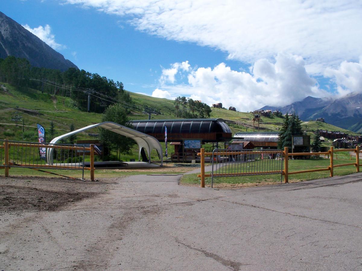 A ski resort entrance featuring a modern chairlift station, surrounded by green hills and mountains under a partly cloudy sky. The area is fenced, with promotional banners visible, indicating recreational activities. Evolution Bike Park at Crested Butte Mountain Resort mountain bike trail.