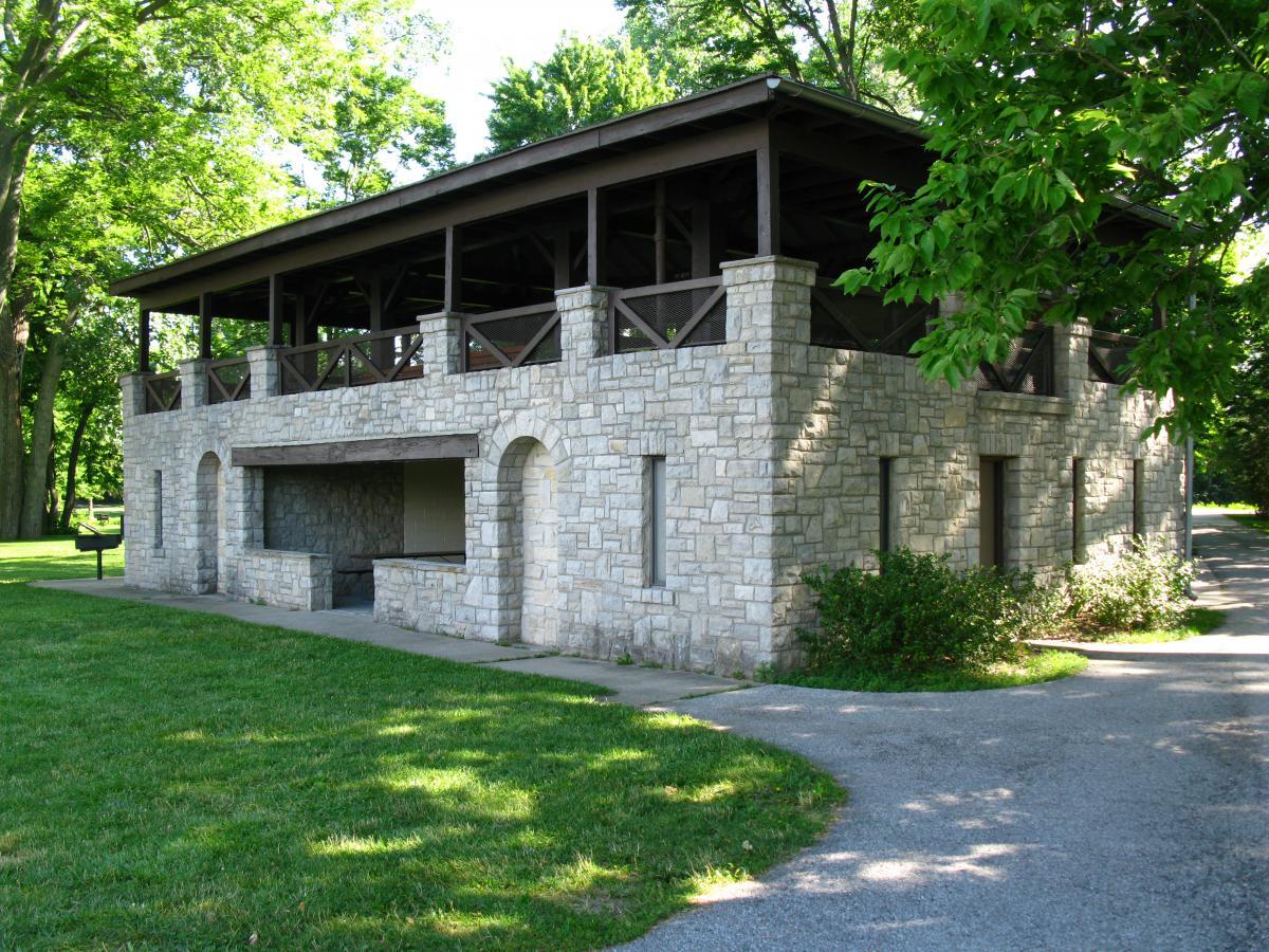 A stone building with a wooden roof and balcony, surrounded by green trees and grass. The structure features arched openings on the lower level and is situated near a pathway, creating a serene outdoor setting. Farnsworth mountain bike trail.