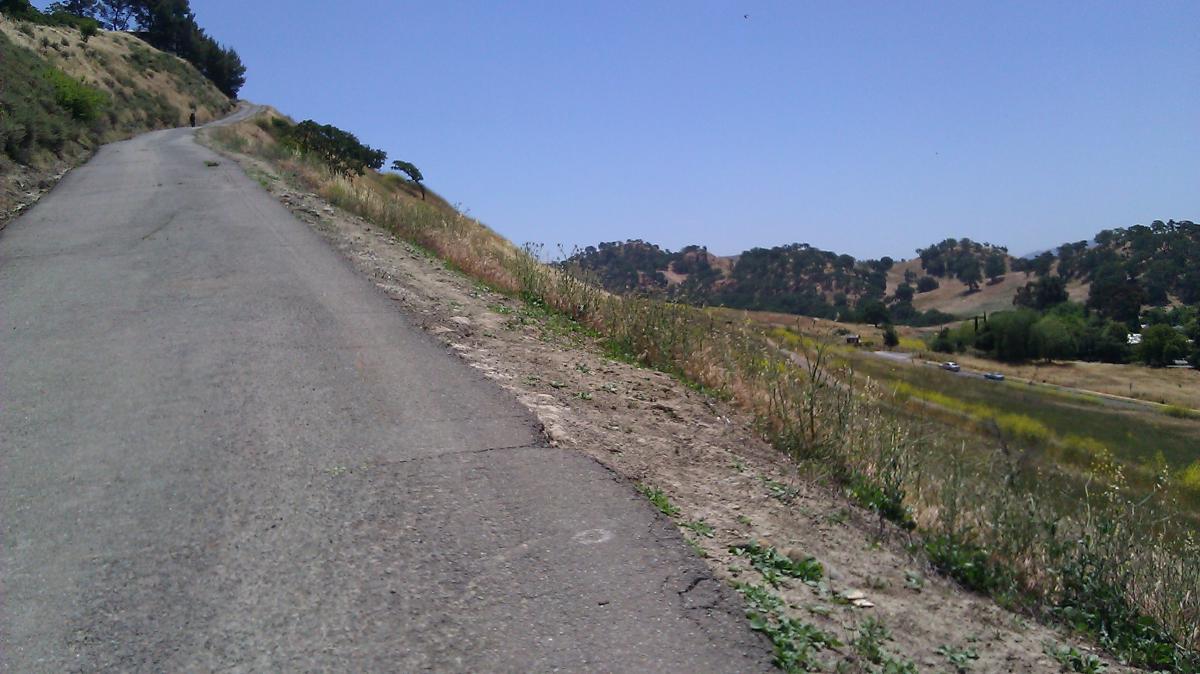 A winding, unpaved road ascending a hillside, surrounded by grassy terrain and sparse vegetation, with rolling hills visible in the background under a clear blue sky. Shell Ridge Open Space mountain bike trail.