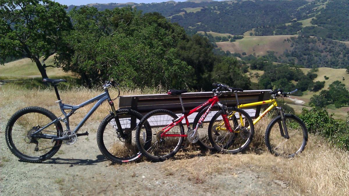 Three mountain bikes—one blue, one red, and one yellow—rest beside a bench on a hillside, with a scenic view of rolling green hills and trees in the background under a clear sky. The ground is dry with patches of grass. Sunol Regional Wilderness mountain bike trail.