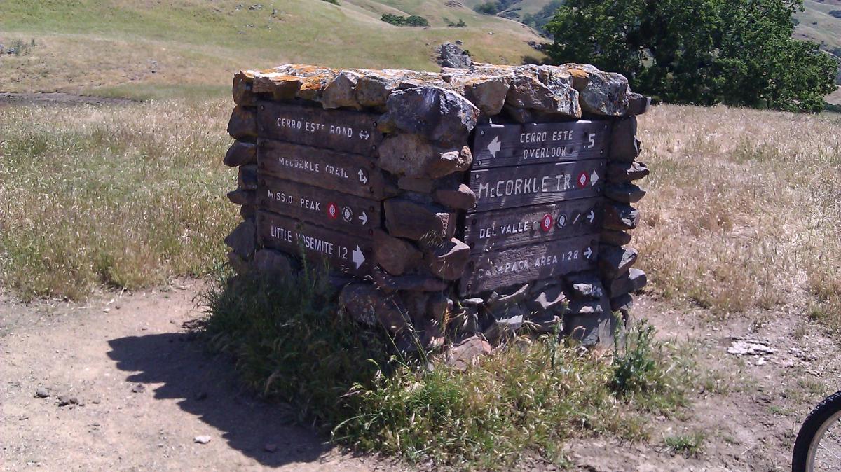 A stone signpost surrounded by grass, displaying directional arrows and distances to various trails and landmarks, including Cerro Este Road, McCorkle Trail, Missio Peak, Little Yosemite, and Del Valle. The sign is weathered and set against a backdrop of rolling hills. Sunol Regional Wilderness mountain bike trail.