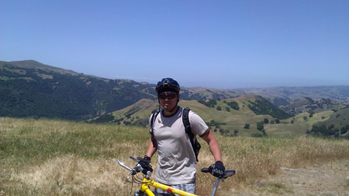 A mountain biker stands on a grassy hilltop, holding a yellow bicycle and wearing a helmet and sunglasses. In the background, expansive green hills stretch out under a clear blue sky. Sunol Regional Wilderness mountain bike trail.