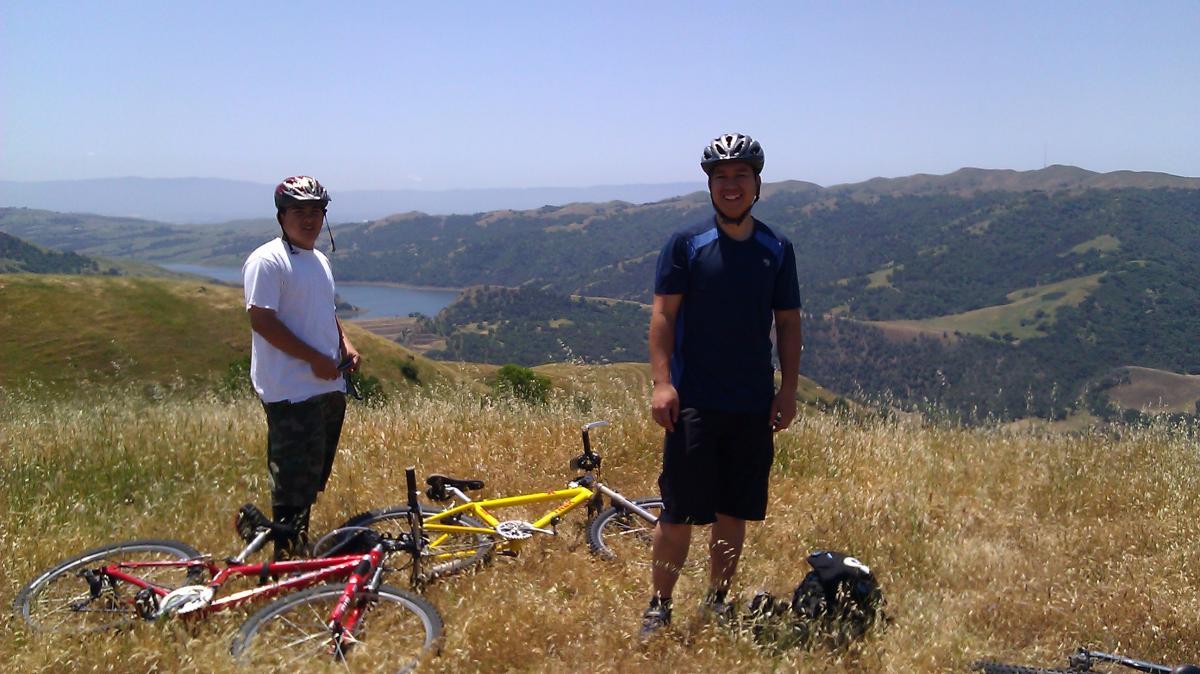 Two cyclists are standing on a grassy hillside with a scenic view of rolling hills and a lake in the background. One cyclist is wearing a white shirt and a helmet, while the other is in a blue shirt. Their bicycles, one red and one yellow, are laid on the ground beside them. The sun is shining, and the landscape is lush and green. Sunol Regional Wilderness mountain bike trail.