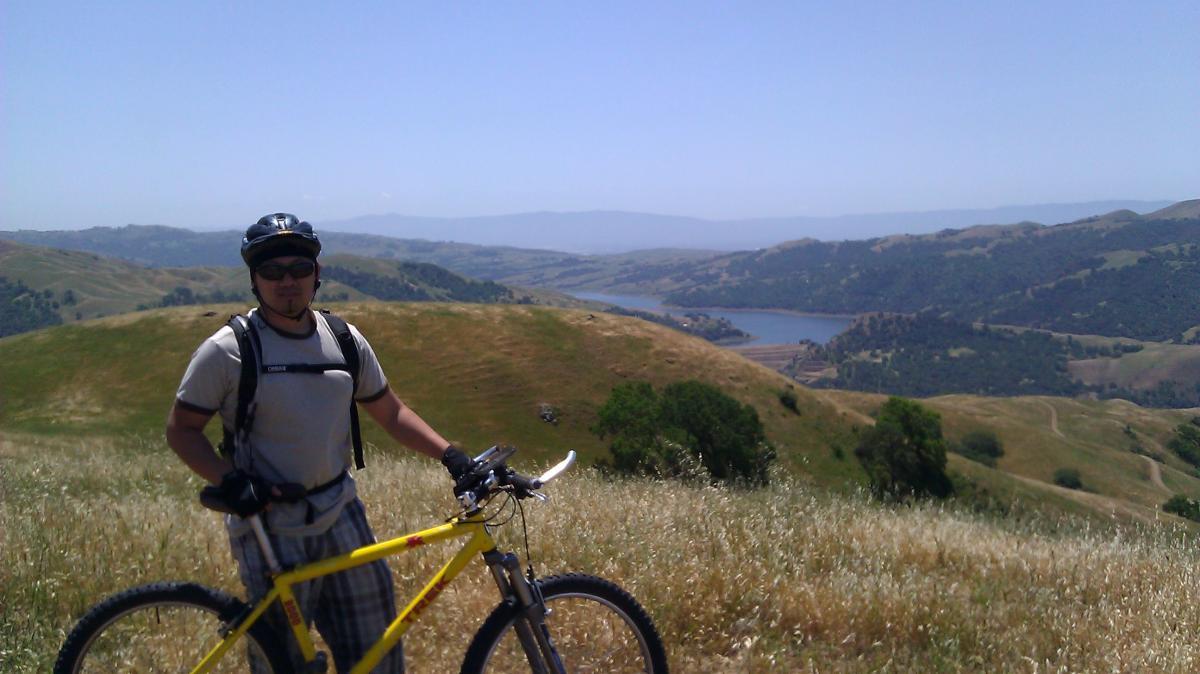 A person wearing a helmet and sunglasses stands next to a yellow mountain bike on a grassy hillside, overlooking a scenic landscape that includes rolling hills and a calm lake in the distance under a clear blue sky. Sunol Regional Wilderness mountain bike trail.