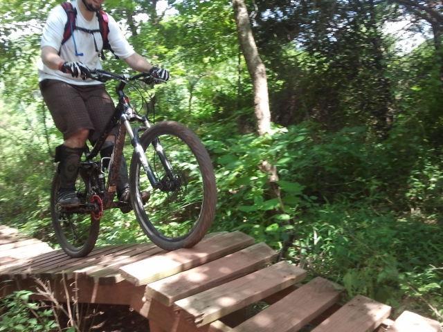 A mountain biker navigating a wooden bridge on a forest trail, surrounded by lush greenery. The rider is mid-jump, with one wheel off the ground, wearing a helmet and casual biking gear. Six Mile Run mountain bike trail.