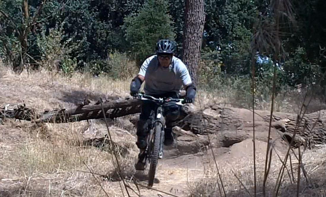 A mountain biker in a grey shirt and black riding gear navigates a dirt trail, maneuvering over a fallen log in a wooded area with greenery in the background. The cyclist is wearing a helmet and gloves, focused on the path ahead. Ripon Riding Spot mountain bike trail.