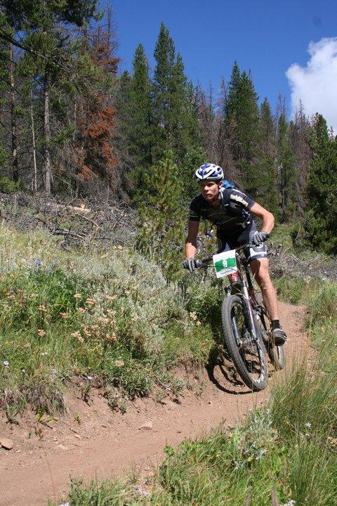 A mountain biker navigating a winding dirt trail in a forested area, surrounded by green vegetation and trees. The cyclist, wearing a helmet and racing attire, appears focused as they ride along the path under a clear blue sky. Colorado Trail mountain bike trail.