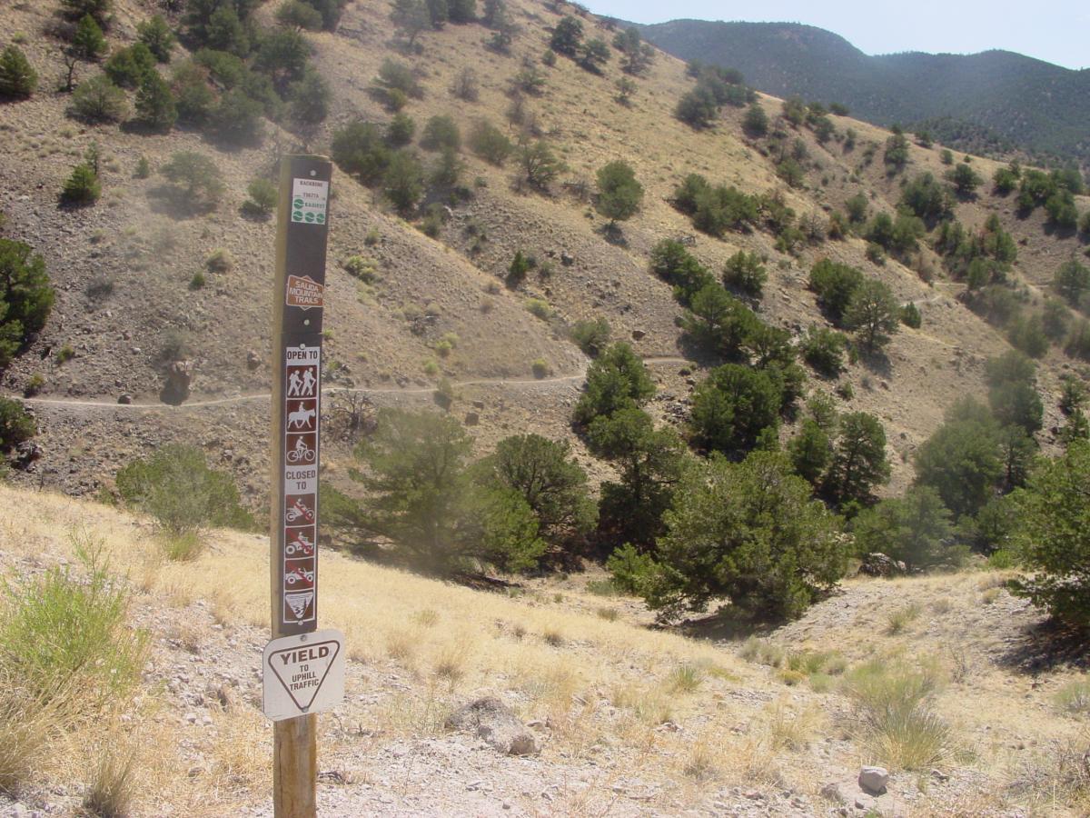 Trail sign indicating usage rules for different types of trail users, including hikers, bicyclists, and horses. The sign features symbols indicating which activities are allowed and lists restrictions for motorized vehicles. The background shows a mountainous landscape with dry grasses and scattered trees. Arkansas Hills mountain bike trail.