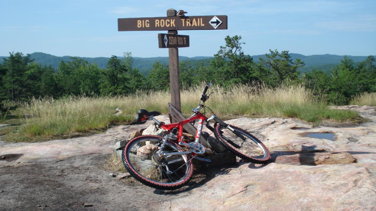 A mountain bike resting on a rocky surface next to a wooden trail sign indicating "Big Rock Trail" and pointing towards "Cedar Rock Trail," surrounded by green trees and rolling hills in the background under a clear blue sky. DuPont State Recreational Forest mountain bike trail.