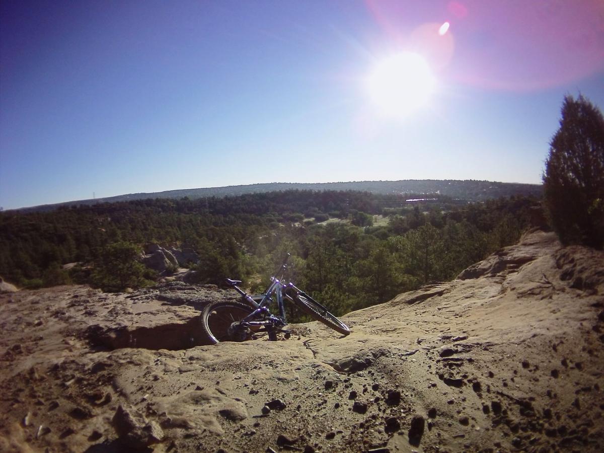 A mountain bike resting on rocky terrain overlooking a vast landscape of trees and hills under a bright sun. Palmer Park mountain bike trail.