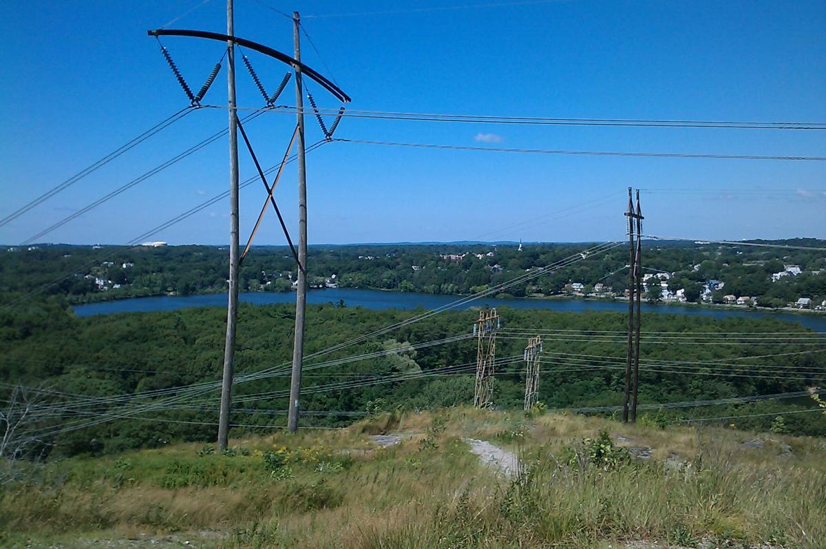 A panoramic view of a landscape featuring tall power lines in the foreground with a tranquil lake and greenery in the background, under a clear blue sky. Horn Pond mountain bike trail.