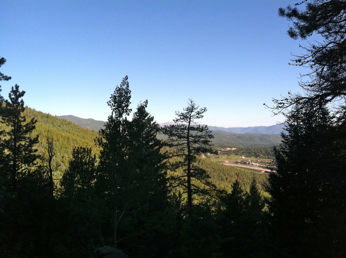 A scenic view of a mountainous landscape with dense green forests, under a clear blue sky. In the foreground, various trees are visible, while the background features rolling hills and distant mountains. A glimpse of a small town is seen nestled in the valley below. Meyer Ranch Park mountain bike trail.