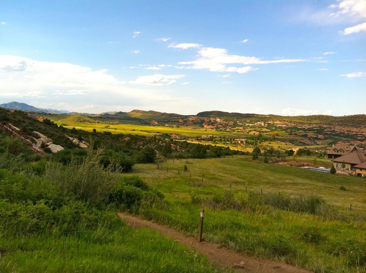 A scenic landscape featuring rolling hills and a vibrant green valley under a blue sky with scattered clouds. In the distance, there are mountains and a few residential buildings nestled among the foliage. A dirt path leads through the foreground, surrounded by lush grass and foliage. Deer Creek Canyon mountain bike trail.